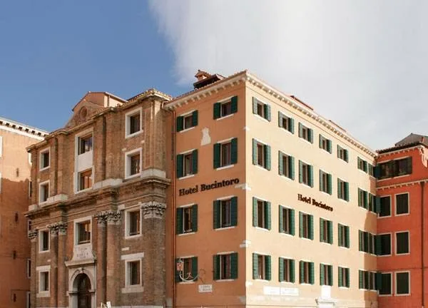 Hotel Bucintoro facade on Riva San Biasio at early morning, pastel stucco, green shutters and the lagoon visible beyond moored gondolas
